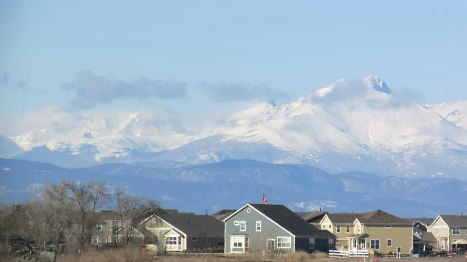 Snow-covered mountains rising above a row of houses on the plain, under a pale blue sky with thin cloud